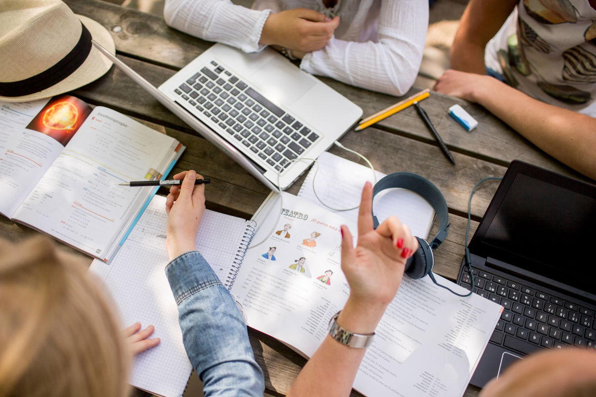 Ungdomar pluggar tillsammans utomhus vid ett bord fullt med datorer och böcker.
